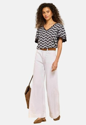 Göndör hajú nő fekete-fehér mintás felsőt, fehér bő szárú nadrágot, barna övet, barna cipőt visel és barna táskát hord.