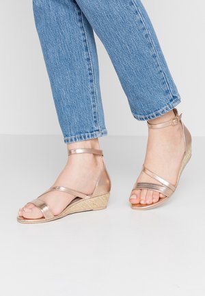 Pieds portant des sandales compensées à bride cheville en or rose associées à un jean en denim bleu, debout sur un sol lisse de couleur claire.