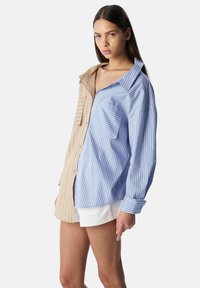 Camicia con bottoni dal design diviso; un lato presenta strisce verticali di un beige chiaro, mentre l'altro è blu con sottili strisce bianche. Vestibilità ampia con grandi tasche.