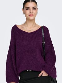 Maglione viola lavorato a maglia con scollo a barca, vestibilità ampia e texture a coste. Indossato sopra una gonna nera, abbinato a una collana e una borsa argentate.