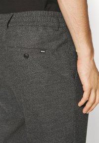 Kolkgrå byxor med mjuk textur, elastisk midjeband, en bakficka med knapp och en liten logotypetikett på fickan.