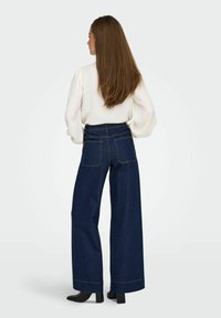Jean en denim bleu foncé à jambes larges et taille haute, associé à un blouse blanche texturée et des bottines à talons noires. Poches arrière visibles.