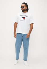 White cotton T-shirt with navy "Tommy Hilfiger" logo and flag graphic. Paired with light blue relaxed-fit jeans and white sneakers.