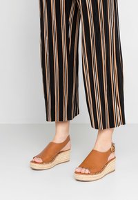 Sandales compensées en cuir tan avec un bout ouvert rond, semelle en jute, et une bride au niveau de la cheville avec une boucle circlaire. Pantalons rayés visibles.