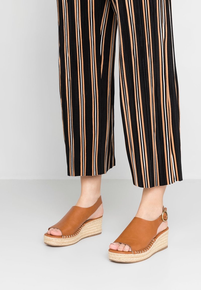 Sandales compensées en cuir tan avec un bout ouvert rond, semelle en jute, et une bride au niveau de la cheville avec une boucle circlaire. Pantalons rayés visibles.