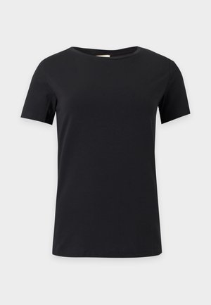 Frau steht mit einer Hand in der Hüfte, trägt ein schlichtes schwarzes T-Shirt, dunkle Jeans mit weitem Bein und helle spitze Schuhe vor weißem Hintergrund.