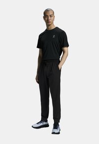 T-shirt noir avec logo sur la poitrine, pantalon de jogging noir avec cordon de serrage, et chaussures de course grises avec accents noirs et semelle à motif.