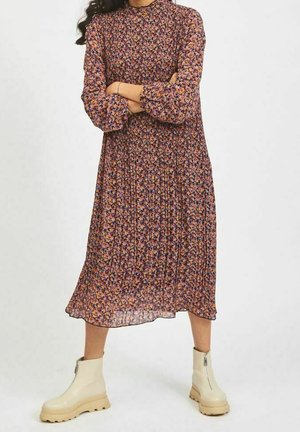 Femme portant une robe midi fleurie à manches longues et col montant, avec des manches bouffantes et des bottines blanches épaisses, debout les bras croisés.