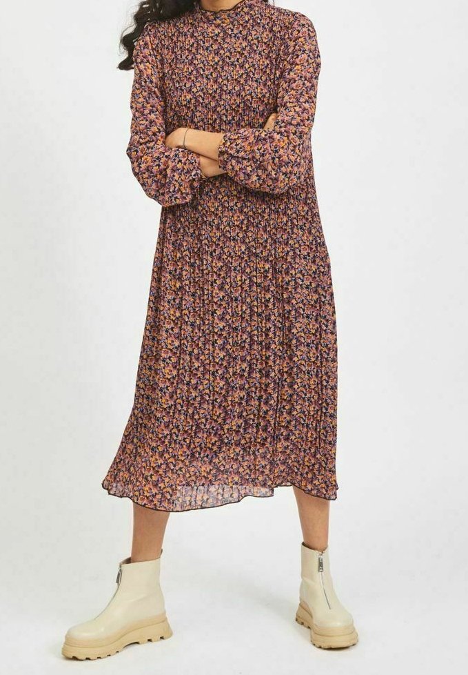 Femme portant une robe midi fleurie à manches longues et col montant, avec des manches bouffantes et des bottines blanches épaisses, debout les bras croisés.