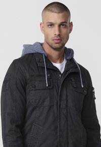 Melna jaka ar pelēku kapuci, kurai ir vairākas kabatas un rāvējslēdzēji. Gluda tekstūra ar ikdienišķu dizainu, piemērota dažādiem laikapstākļiem.