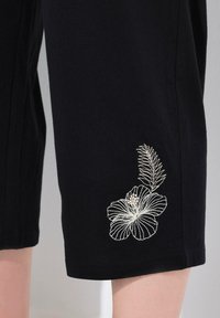 Pantalones de algodón negros con bordado floral blanco en la parte inferior de la pierna izquierda. La tela es suave, con un ajuste relajado y un corte recto.