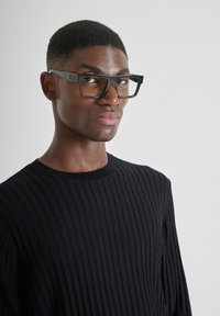 Occhiali neri rettangolari con montatura spessa, indossati sopra un maglione a coste nero e testurizzato. Gli occhiali presentano un discreto dettaglio del marchio sulla montatura.
