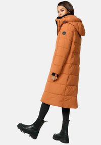 Abrigo largo acolchado en naranja, con capucha desmontable, cierre frontal con cremallera y bolsillos laterales. Combinado con jeans ajustados negros y botas negras gruesas.