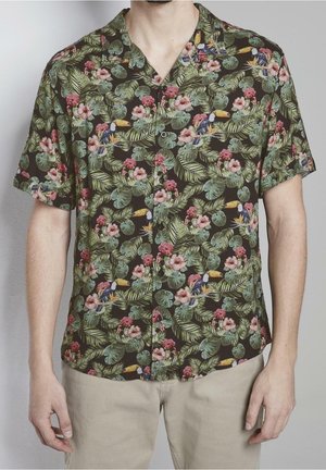Homme portant une chemise à manches courtes boutonnée avec un imprimé de feuilles tropicales vertes, de fleurs roses et de toucan, associée à un pantalon beige.