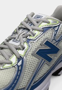 Κοντινό πλάνο ενός αθλητικού παπουτσιού από γκρι και μπλε δίχτυ, με ανοιχτογκρι κορδόνια και νέον πράσινες λεπτομέρειες στο πάνω μέρος.