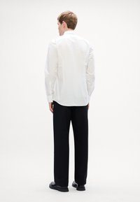 Witte blouse met lange mouwen en een knoopsluiting op de rug, gecombineerd met zwarte broek en zwarte schoenen. Simpele, gladde textuur, klassieke pasvorm.