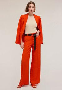 Blazer su misura arancione brillante abbinato a pantaloni larghi arancioni, con una cintura nera. Il modello indossa un top a coste crema e sandali arancioni.