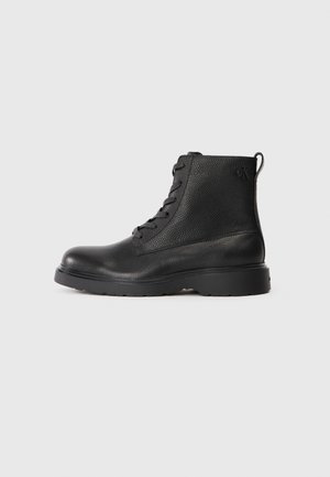 Black ankle boot made of textured leather with a round toe, lace-up design, and a sturdy black rubber sole. Features a loop at the heel.