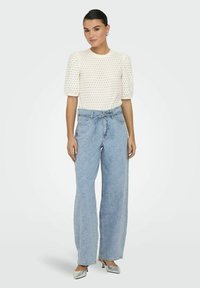 Haut crème léger texturé avec des manches bouffantes et des pois, associé à un jean large à taille haute, à motif floral, bleu clair, et des escarpins argentés.