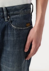 Jeans de ganga azul escura com uma textura desgastada, apresentando um rótulo G-Star Originals no bolso da frente e um botão de metal na cintura.