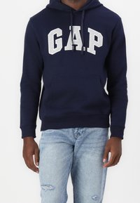 Sudadera azul marino con el logo "GAP" en gris claro. Presenta un bolsillo frontal, capucha con cordón y puños acanalados. Combinada con jeans azul claros.