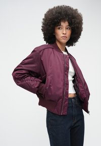 Cazadora bomber burdeos con acabado satinado suave, puños acanalados y cremallera frontal, combinada con vaqueros denim oscuros y una camiseta blanca.