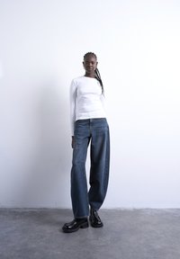 Νέο άτομο με πλεγμένα μαλλιά, που φοράει λευκό μακρυμάνικο τοπ, φαρδιά μπλε τζιν και μαύρα παπούτσια με πάτο, μπροστά σε απλό τοίχο.