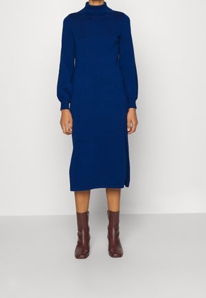 Person trägt ein blaues, langärmliges, knielanges Kleid und braune Stiefeletten und steht vor einem schlichten weißen Hintergrund.