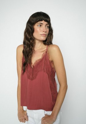 Woman with long wavy dark hair wearing a sleeveless rust-colored top with lace details, looking to her right against a plain light background.