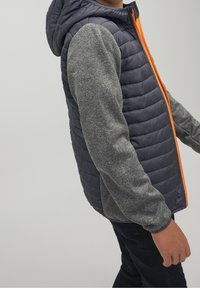 Padded jacket w niebieskiej korpusie, szarych rękawach i pomarańczowym zamku. Posiada kaptur oraz strukturalną tkaninę dla zapewnienia ciepła.