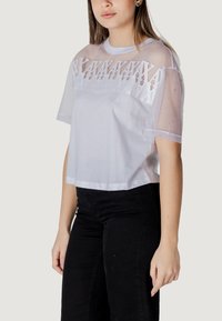 Camiseta corta blanca con mangas de malla transparente y detalles bordados en el pecho. La tela es suave, con un corte relajado y escote redondo.