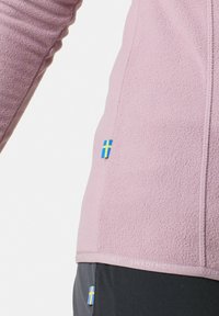 Giacca in pile di colore rosa chiaro, con texture morbida, dotata di chiusura con zip e dettagli ricamati della bandiera svedese sull'orlo e sulla manica.
