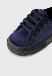 Sneaker albastru din suede cu o talpă din cauciuc texturat negru, având șase șireturi negre trecute prin ochiuri circulare.