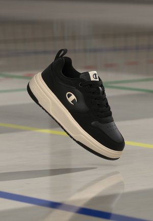 Ασπρόμαυρο sneaker χαμηλό με κορδόνια και λογότυπο στο πλάι και στη γλώσσα, εμφανιζόμενο στον αέρα πάνω από πάτωμα γηπέδου αθλοπαιδιών.