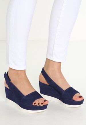 Navy blue suede wedge sandals with open toes and slingback design, featuring a white textured sole and minimalistic accents.