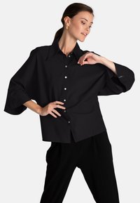 Camicia nera oversize con colletto appuntito, maniche lunghe e bottoni argento lungo la parte anteriore. Realizzata in tessuto liscio con una vestibilità comoda.