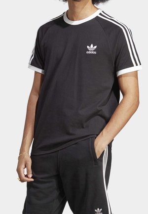 Mężczyzna w czarnej koszulce Adidas i dopasowanych spodniach z białymi paskami na rękawach i bokach, jedna ręka w kieszeni.