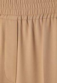 Beige rekbare stof met een gestructureerd oppervlak, voorzien van een elastische tailleband en een zijzakdetail. Soepel, flexibel materiaal.