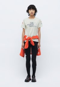 T-shirt gris clair à manches courtes avec le logo "The North Face", leggings noirs, et une veste orange nouée autour de la taille. Chaussures noires.