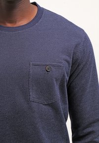 Navy long-sleeve shirt made of textured fabric, featuring a chest pocket with a round black button and small white dot pattern throughout.