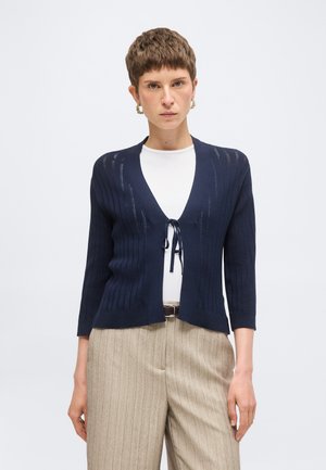 Kobieta w granatowym dzianinowym kardiganie zawiązanym z przodu, białym topie, beżowych spodniach w prążki i złotych kolczykach-kołach, stojąca na jednolitym tle.