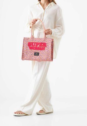 Personne vêtue de blanc tenant un sac fourre-tout imprimé léopard rose et orange avec l'inscription "SUPER WOMAN" en lettres rouges épaisses.