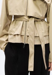 Μπεζ σακάκι με ζώνη και φερμουάρ μπροστά, φορεμένο πάνω από μαύρο παντελόνι με λεπτές λευκές ρίγες, εμφανίζοντας κοντινή λήψη της μέσης και του κορμού.