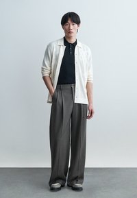 Jeune homme debout les mains dans les poches, portant un cardigan crème clair boutonné sur une chemise noire, un pantalon gris à jambes larges et des chaussures bicolores.