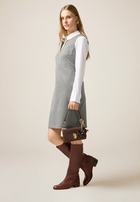 Grau-weißes Hahnentrittkleid mit einem Kragenhemd darunter, kombiniert mit braunen kniehohen Stiefeln und einer braunen strukturierten Handtasche.