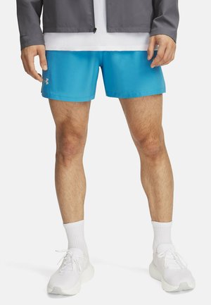 Pantalones cortos deportivos azules con una textura suave, logo en el lado izquierdo, combinados con zapatillas y calcetines blancos. Vello en las piernas visible y piernas tonificadas.
