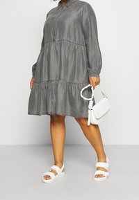 Grijze, lange mouwen tiered jurk met kraag; gecombineerd met een witte schoudertas en witte sandalen. De jurk heeft een zachte, gestructureerde stof.