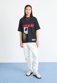 Black oversized t-shirt featuring bold red and yellow graphic prints, paired with white sweatpants and gray-green sneakers.