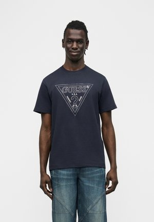 Muž stojaci oblečený v čiernom tričku s logom GUESS a modrých džínsach na bielom pozadí, otočený čelom so slabým úsmevom.