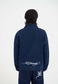 Námořnická džínová bunda s výrazným vyšitým logem Ed Hardy v bílé barvě na zádech. Disponuje klasickým límcem a volným střihem.